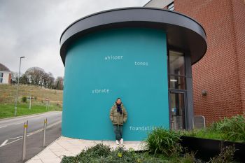 Helen Cammock stands in front of the artwork on an external wall at top of 海角社区 Student Centre. The text is painted in white and set against a bright teal background, and reads: whisper  tones  vibrate  foundations