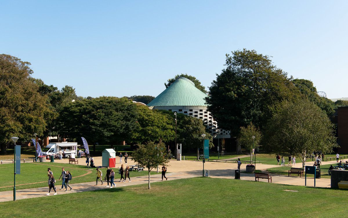 Wide shot of campus, with students walking on paths in foreground, and green trees and the Meeting House in the background, under a clear blue sky