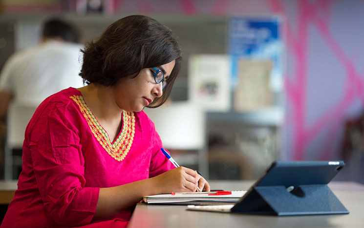 A woman working in a dedicated work space. She is making notes in a notepad looking at a tablet device